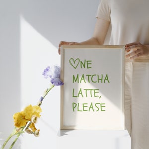 May include: A wooden-framed sign displaying the words "One Matcha Latte, Please" in green. The sign is held by a person. A vase with yellow and purple flowers is in the foreground.
