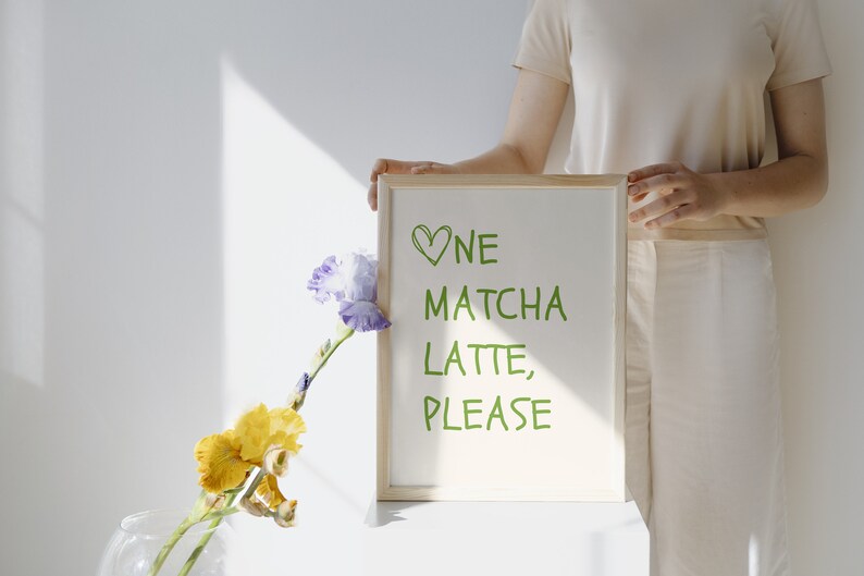May include: A wooden-framed sign displaying the words "One Matcha Latte, Please" in green. The sign is held by a person. A vase with yellow and purple flowers is in the foreground.