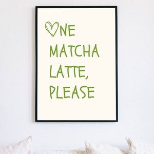 May include: A framed print featuring the text "One Matcha Latte, Please" in green, handwritten lettering. The words are set against a cream backdrop, with a green heart above "One". The frame is black.