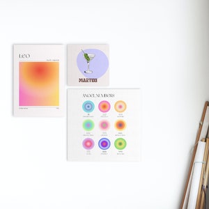 May include: Three colourful prints on a white wall. The first print is a gradient of pink, orange, and yellow with the word "LEO" at the top. The second print is a light blue background with a martini glass and the word "MARTINI" at the bottom. The third print is a light green background with the words "ANGEL NUMBERS" at the top and nine colourful circles with numbers inside.