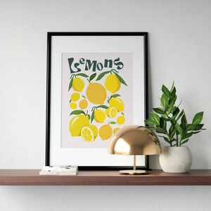 May include: A framed print with a light pink background and the text "Les Mons" in green. The print features a cluster of yellow lemons with green leaves.