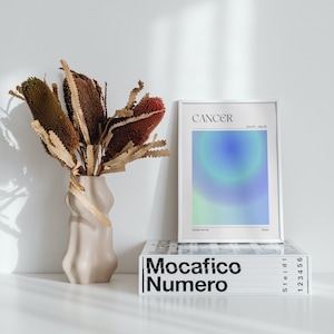 May include: A white framed print with the word "Cancer" and a blue and green gradient design. The print is sitting on top of a book titled "Mocacifico Numero" with black text on a white background.