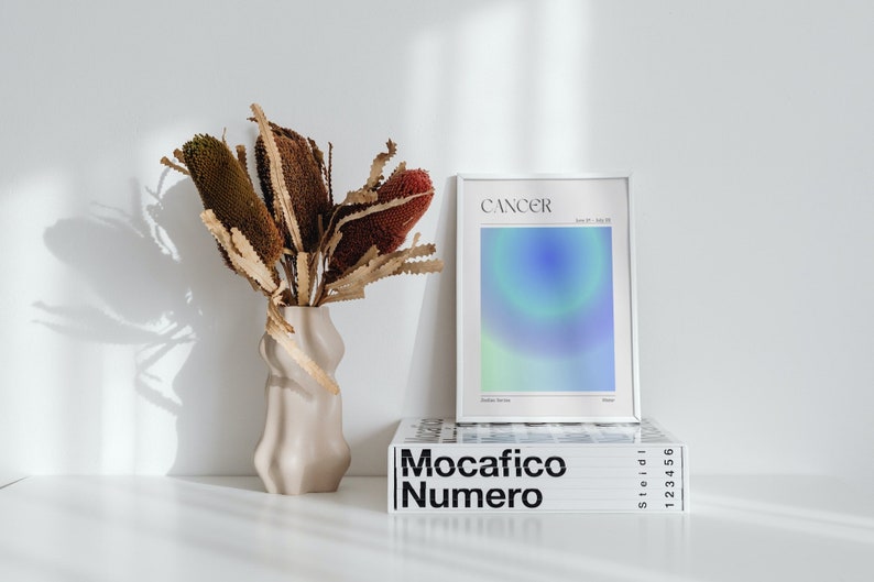 May include: A white framed print with the word "Cancer" and a blue and green gradient design. The print is sitting on top of a book titled "Mocacifico Numero" with black text on a white background.