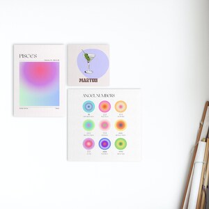 May include: Three colorful prints on a white wall. The first print is a gradient of pink, blue, and green with the word "Pisces" at the top. The second print is a light blue background with a martini glass and the word "Martini" at the bottom. The third print is a light green background with the words "Angel Numbers" at the top and nine colorful circles with numbers inside.