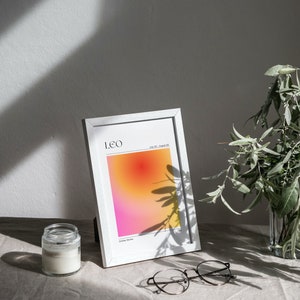May include: A white frame with a print of a gradient background in shades of pink, orange, and yellow. The print has the text "LEO" and "July 23 - August 22" in black. The frame is sitting on a light brown surface with a glass jar and round glasses.