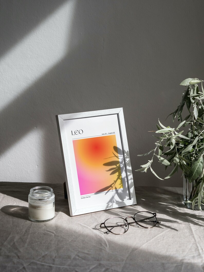 May include: A white frame with a print of a gradient background in shades of pink, orange, and yellow. The print has the text "LEO" and "July 23 - August 22" in black. The frame is sitting on a light brown surface with a glass jar and round glasses.