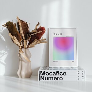 May include: A white framed print with the word "Pisces" and a gradient of pink, purple, green, and blue colors. The print is sitting on top of a stack of books with the title "Mocacifico Numero" in black text.