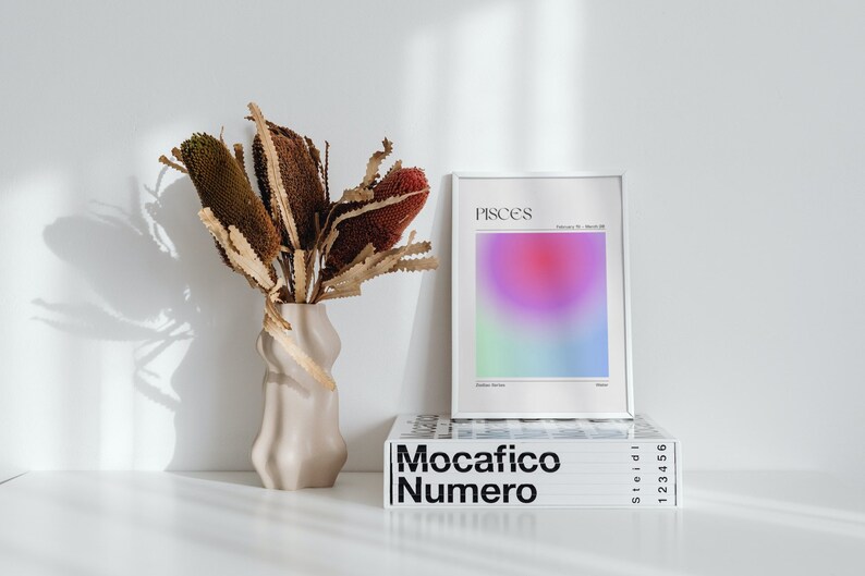 May include: A white framed print with the word "Pisces" and a gradient of pink, purple, green, and blue colors. The print is sitting on top of a stack of books with the title "Mocacifico Numero" in black text.