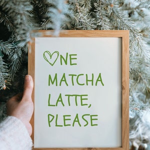 May include: A wooden-framed sign with the text "One Matcha Latte, Please" in green lettering. A green heart is drawn above the word "One". The sign is held in front of a backdrop of green pine needles.