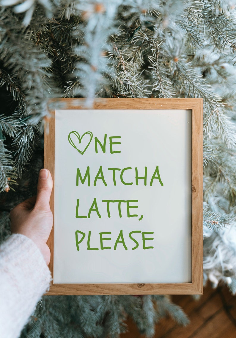 May include: A wooden-framed sign with the text "One Matcha Latte, Please" in green lettering. A green heart is drawn above the word "One". The sign is held in front of a backdrop of green pine needles.