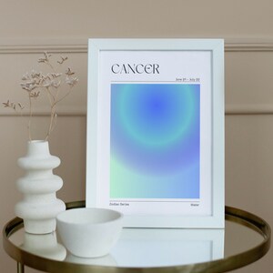 May include: A white framed print with the word "CANCER" in black text at the top. The print features a blue, green, and white gradient background with a circle in the center. The text "June 21 - July 22" is below the word "CANCER". The text "Zodiac Series" is in the bottom left corner and the text "Water" is in the bottom right corner.