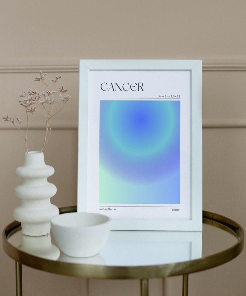 May include: A white framed print with the word "CANCER" in black text at the top. The print features a blue, green, and white gradient background with a circle in the center. The text "June 21 - July 22" is below the word "CANCER". The text "Zodiac Series" is in the bottom left corner and the text "Water" is in the bottom right corner.