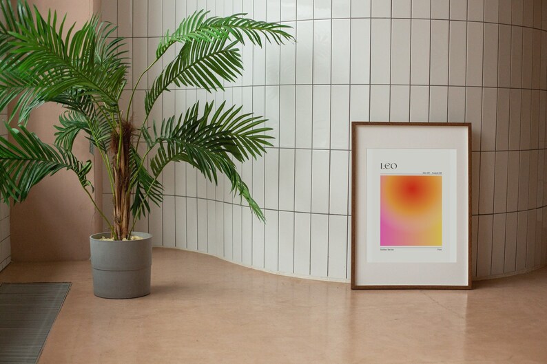 May include: A framed print with a gradient background in shades of pink, orange, and yellow. The print has the word "LEO" in black text at the top and the dates "July 23 - August 22" in smaller black text below. The frame is brown wood.
