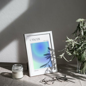 May include: A white framed print with a blue and green gradient background. The print says "Cancer" and "June 21 - July 22" in black text. The print is sitting on a light brown surface with a glass jar and glasses.