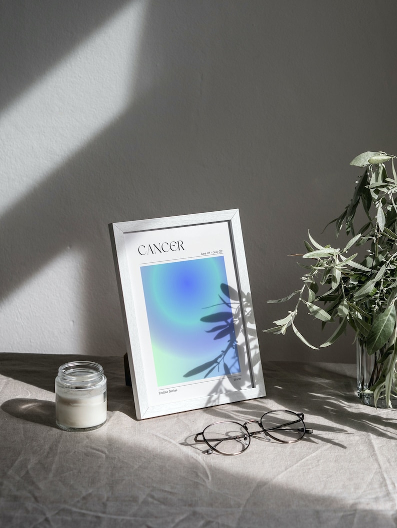 May include: A white framed print with a blue and green gradient background. The print says "Cancer" and "June 21 - July 22" in black text. The print is sitting on a light brown surface with a glass jar and glasses.