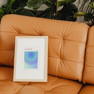 May include: A framed print with a light blue, green, and purple gradient design. The print has the word "CANCER" and the dates "June 21 - July 22" printed on it. The print is sitting on a brown leather couch.