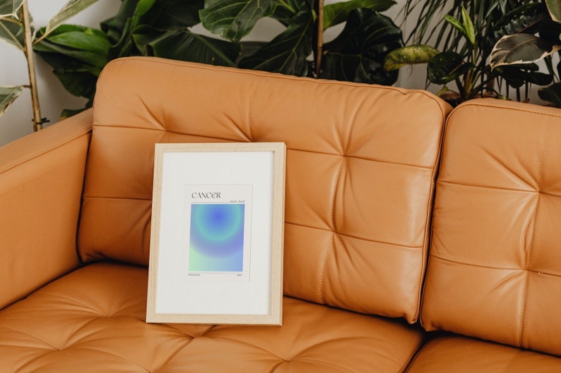 May include: A framed print with a light blue, green, and purple gradient design. The print has the word "CANCER" and the dates "June 21 - July 22" printed on it. The print is sitting on a brown leather couch.