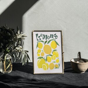 May include: A framed print with a yellow and green illustration of lemons and the word "Lemons" in a retro font. The print is sitting on a black surface with a small bowl and a spoon.