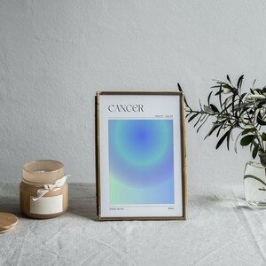 May include: A gold framed print with a blue and green gradient design. The print has the word "Cancer" and the dates "June 21 - July 22" printed on it. The print is sitting on a white table cloth with a candle and a vase of greenery.