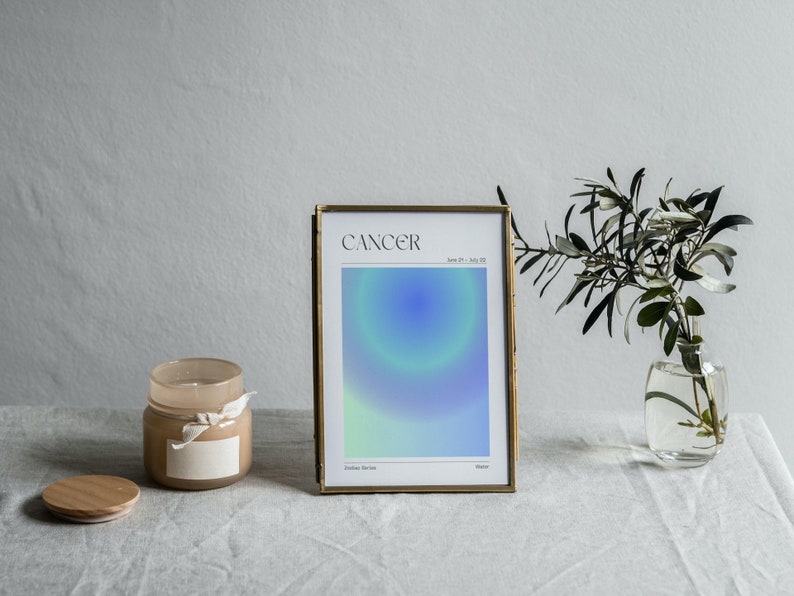 May include: A gold framed print with a blue and green gradient design. The print has the word "Cancer" and the dates "June 21 - July 22" printed on it. The print is sitting on a white table cloth with a candle and a vase of greenery.