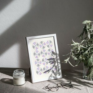 K&ouml;nnte beinhalten: Ein wei&szlig;er Bilderrahmen mit silbernem Rand zeigt ein Raster aus 16 silbernen CDs. Der Rahmen steht auf einer hellbraunen Oberfl&auml;che mit einem durchsichtigen Glasgef&auml;&szlig; und einer Brille.