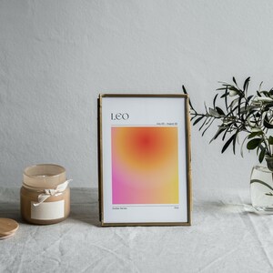 May include: A gold framed print with a gradient background in shades of pink, orange, and yellow. The print has the text "LEO" and "July 23 - August 22" in black. The print is sitting on a white tablecloth with a candle and a vase of greenery.