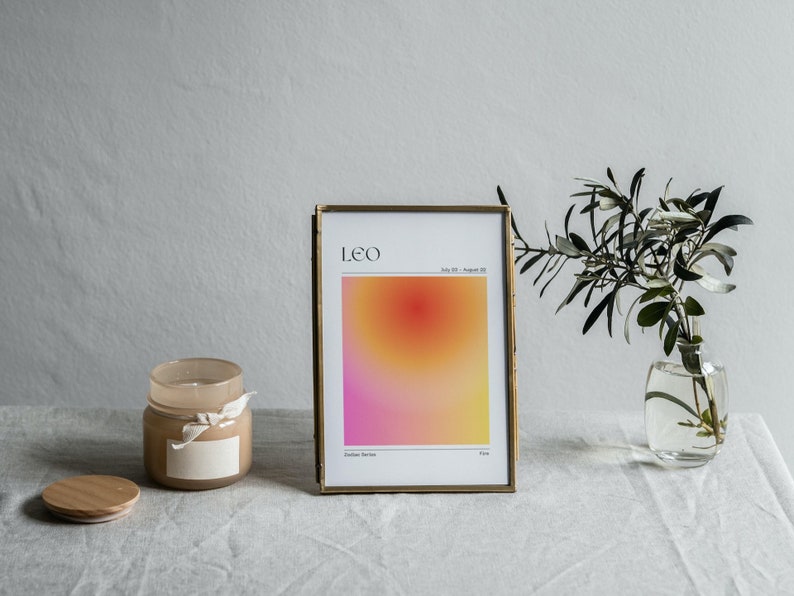 May include: A gold framed print with a gradient background in shades of pink, orange, and yellow. The print has the text "LEO" and "July 23 - August 22" in black. The print is sitting on a white tablecloth with a candle and a vase of greenery.