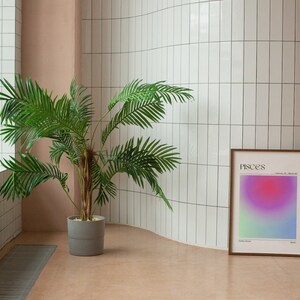 May include: A framed print with a gradient background in shades of pink, purple, and green. The print has the word "Pisces" and the dates "February 19 - March 20" printed in black text. The print is hanging on a white tiled wall next to a potted palm tree.