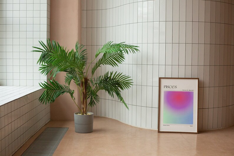 May include: A framed print with a gradient background in shades of pink, purple, and green. The print has the word "Pisces" and the dates "February 19 - March 20" printed in black text. The print is hanging on a white tiled wall next to a potted palm tree.