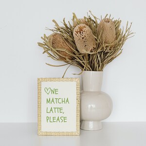 May include: A beige vase displays dried flowers, beside a framed sign. The sign has a woven border and green text that reads "One Matcha Latte, Please" with a heart symbol. The vase and sign are on a white surface.