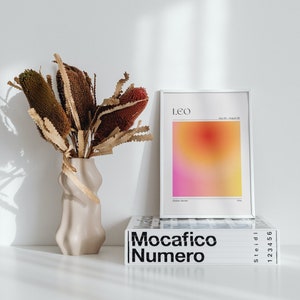 May include: A white framed print with a gradient background in shades of pink, orange, and yellow. The print has the word "Leo" in black text at the top and the text "July 23 - August 22" in smaller black text below. The print is sitting on top of a white book with the text "Mocacifico Numero" in black text.