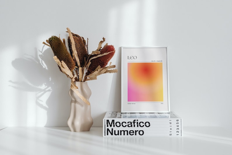 May include: A white framed print with a gradient background in shades of pink, orange, and yellow. The print has the word "Leo" in black text at the top and the text "July 23 - August 22" in smaller black text below. The print is sitting on top of a white book with the text "Mocacifico Numero" in black text.