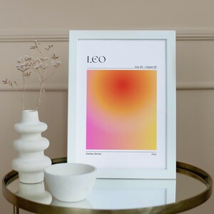 May include: White framed print with a gradient background in shades of pink, orange, and yellow. The print features the word "LEO" in black text at the top and the dates "July 23 - August 22" in smaller black text below. The words "Zodiac Series" and "Fire" are printed in black text at the bottom of the print.
