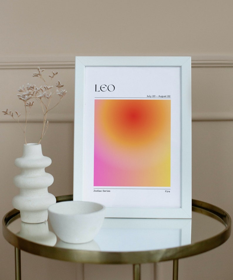 May include: White framed print with a gradient background in shades of pink, orange, and yellow. The print features the word "LEO" in black text at the top and the dates "July 23 - August 22" in smaller black text below. The words "Zodiac Series" and "Fire" are printed in black text at the bottom of the print.