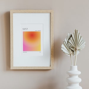May include: A framed print with a gradient background in shades of pink, orange, and yellow. The print features the word "LEO" and the dates "July 23 - August 22". The print is in a light wood frame and is hanging on a light beige wall.