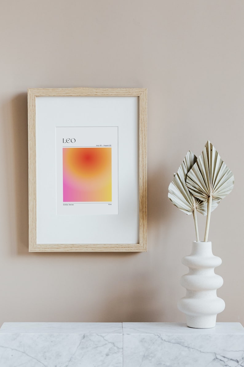 May include: A framed print with a gradient background in shades of pink, orange, and yellow. The print features the word "LEO" and the dates "July 23 - August 22". The print is in a light wood frame and is hanging on a light beige wall.