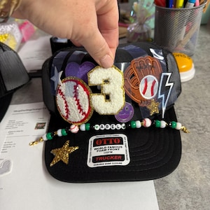 May include: Black trucker hat with baseball-themed patches, including a baseball, glove, and the number "3". The hat features a beaded brim and a gold star. The text "OTTO" and "TRUCKER" are visible.