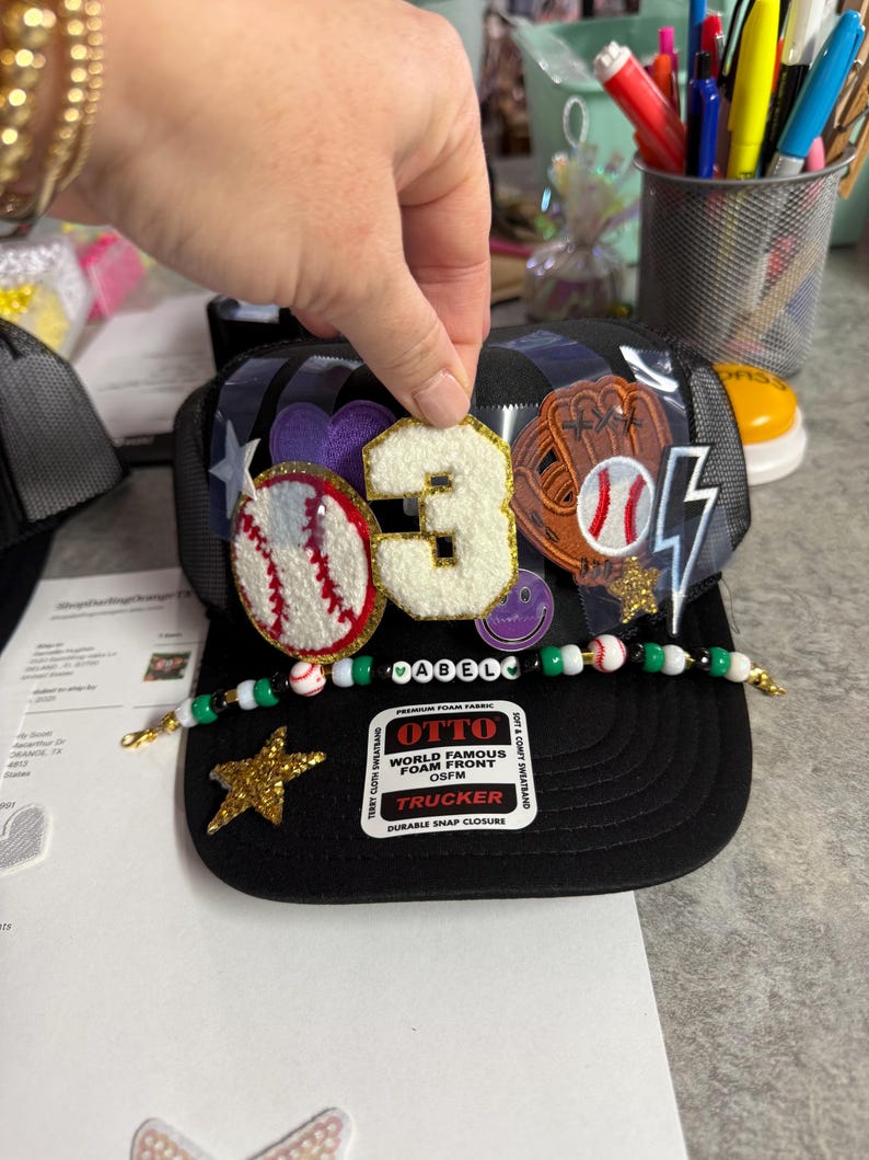 May include: Black trucker hat with baseball-themed patches, including a baseball, glove, and the number "3". The hat features a beaded brim and a gold star. The text "OTTO" and "TRUCKER" are visible.