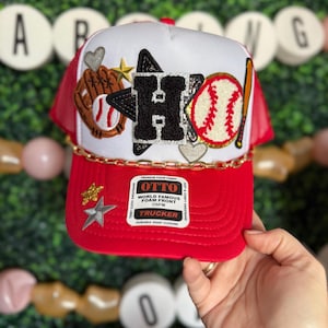 May include: Red and white trucker hat with a black letter H, a baseball, a baseball glove, and a star. The hat has a gold chain and a label that says "OTTO WORLD FAMOUS FOAM FRONT OSFM TRUCKER".