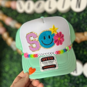 May include: A light green trucker hat with a white front panel. The front panel features a pink and white felt letter "S", a blue smiley face with a gold outline, a pink flower, and a yellow star. The hat has a rainbow beaded trim and a red heart on the side. The hat has a label that reads "Sutton Otto World Famous Foam Front Young Adult Trucker".