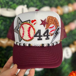 May include: A baseball-themed trucker hat with a white front panel and a maroon brim and mesh back. Embellishments include a baseball, a glove, a star, the number 44, and the name "JACK" spelled out in beads. A heart with the word "MOM" is also present.