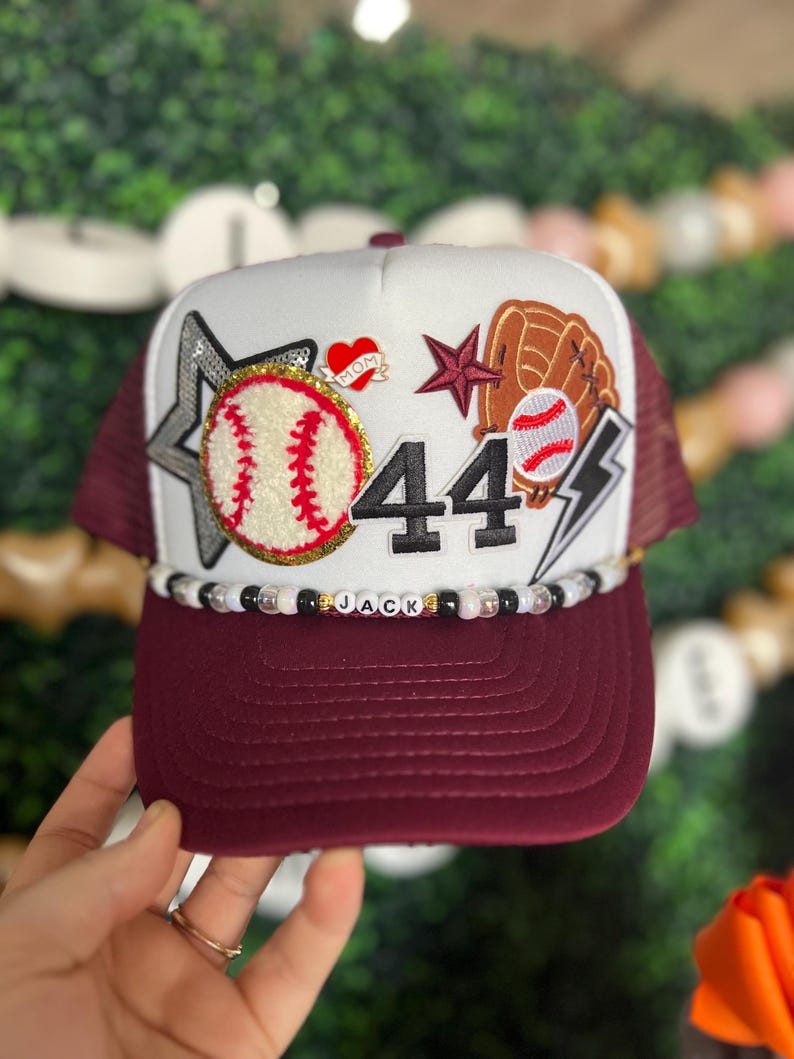 May include: A baseball-themed trucker hat with a white front panel and a maroon brim and mesh back. Embellishments include a baseball, a glove, a star, the number 44, and the name "JACK" spelled out in beads. A heart with the word "MOM" is also present.