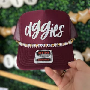 May include: Burgundy trucker hat with a white glitter design that says "doggies" and a gold and white beaded trim. The hat has a label that says "Otto World Famous Foam Front Trucker".