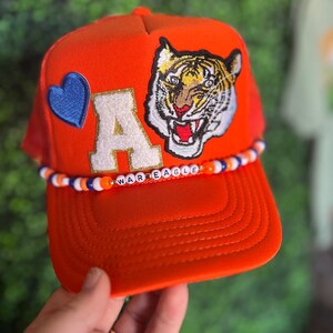 May include: Orange trucker hat with a blue heart, a white letter A with gold glitter, and a tiger face patch. The hat has a beaded band with the words "WAR EAGLE" in white letters.
