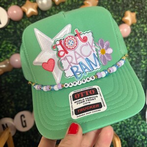 May include: A green trucker hat with a white star, pink heart, and a floral design. The hat features the text "Hot Crack Bam Mahjong" and a beaded band. The hat is made of foam fabric and has a snap closure.