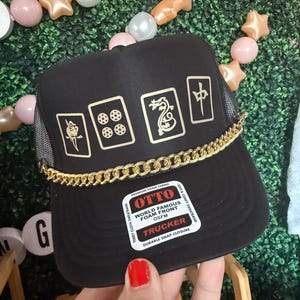 May include: Black trucker hat with gold chain and gold graphic designs. The hat features a mesh back and a curved brim. The front of the hat has a label that reads "OTTO WORLD FAMOUS FOAM FRONT OSFM TRUCKER".