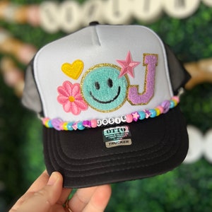 May include: White trucker hat with a black mesh back. The front of the hat features a pink flower, a yellow heart, a turquoise smiley face, a purple letter J with gold glitter, and a pink star. The hat has a beaded trim with the name "JOSIE" spelled out in colorful beads. The hat also has a label that reads "OTTO WORLD FAMOUS FOAM FRONT YOUTH TRUCKER".