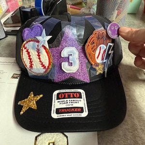 May include: Black trucker hat with a baseball theme. The hat features a glittery purple star with the number 3, a baseball, and a baseball glove. A gold star and a smiley face sticker are also visible.