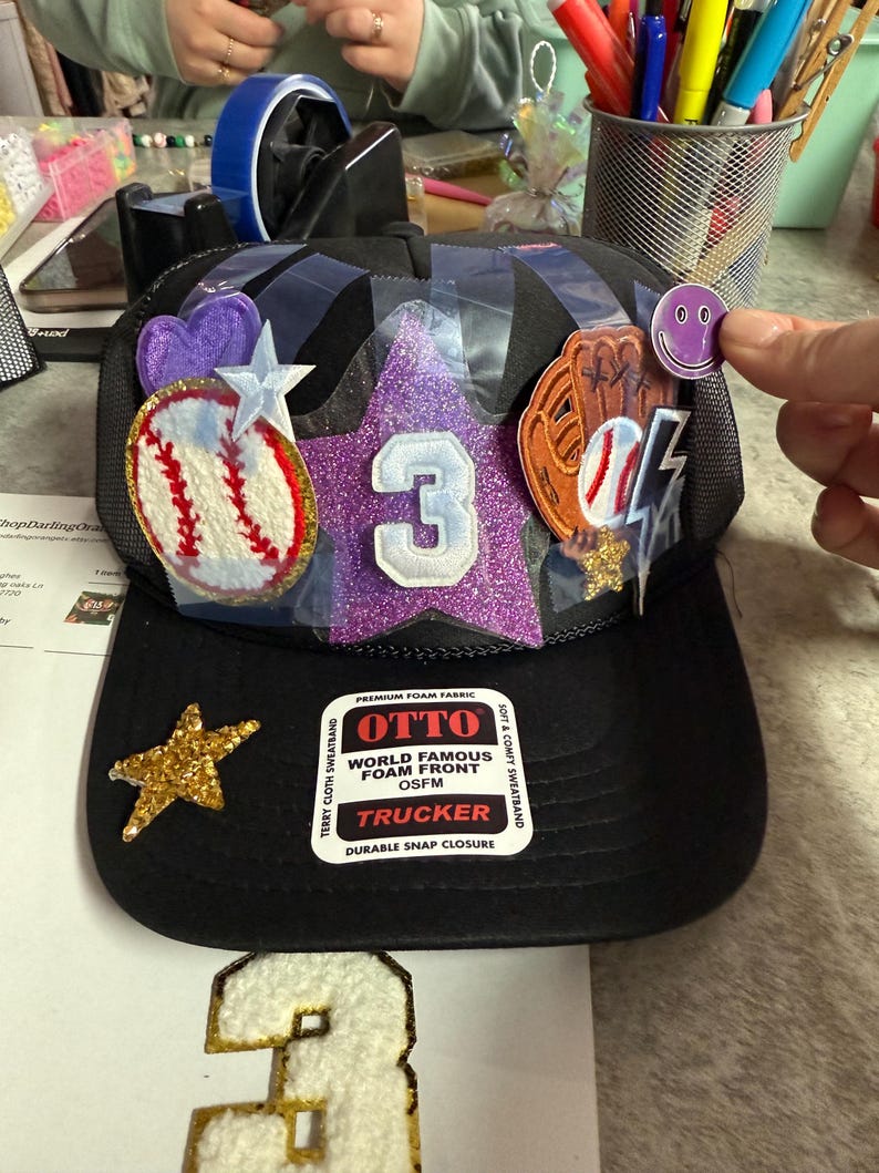 May include: Black trucker hat with a baseball theme. The hat features a glittery purple star with the number 3, a baseball, and a baseball glove. A gold star and a smiley face sticker are also visible.