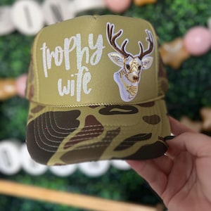 Peut inclure: Un chapeau de camionneur camouflage avec un cerf blanc et le texte "trophy wife" en paillettes blanches.